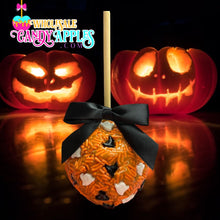 A Halloween-themed candy apple with a black ribbon and white and orange candy decorations sits in front of two carved jack-o-lanterns with glowing orange and red faces.