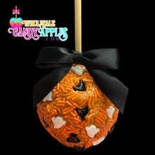 A candy apple with a black ribbon and orange candy coating, decorated with white and black candy pieces, is displayed against a black background.