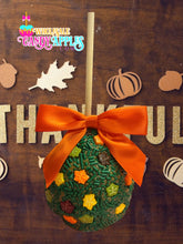 A green candy apple with orange and yellow sprinkles, tied with an orange ribbon, is displayed against a wooden background with autumn-themed decorations.